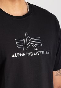 Schwarzes Baumwoll-T-Shirt mit einem metallischen, reflektierenden Logo, das ein stilisiertes "A" und horizontale Linien zeigt. Rundhalsausschnitt und kurze Ärmel.