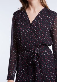 Zwarte jurk met een V-hals, knoopsluiting en lange doorzichtige mouwen met een klein hartjespatroon in rood, wit en blauw. Vastgebonden in de taille.