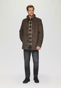 Bruine wolmix jas met een kraag, zwarte accenten en grote knopen; gelaagd over een zwart-beige geruit overhemd, gecombineerd met donkere jeans.