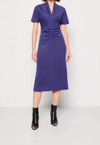 Fialové šaty s krátkým rukávem a výstřihem do V, s nařasením v pase a rovnou midi délkou. Nosené s černými kotníkovými botami.