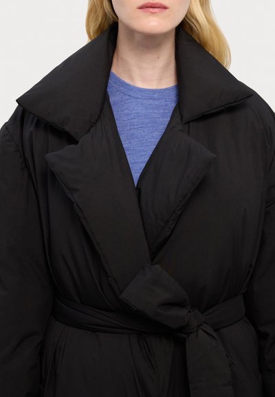 Black padded coat with large collar and tie waist. Soft texture, oversized fit, stitched detailing, worn over a blue long-sleeve shirt.