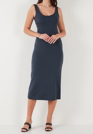 Kobieta ubrana w granatową, dopasowaną sukienkę na ramiączkach o długości midi, czarne sandały na słupku z odkrytymi palcami, srebrny naszyjnik i bransoletkę.