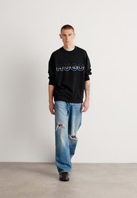 Felpa nera con logo "HUGO" in colori a contrasto, abbinata a jeans blu strappati con buchi e calzature nere. Sfondo semplice.