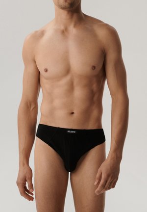 Mężczyzna bez koszulki w czarnych slipach marki Atlantic, stojący z rozluźnionymi ramionami na jasnym, jednolitym tle.