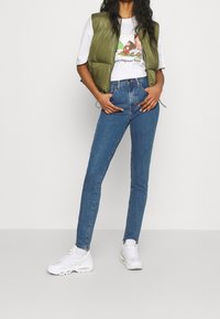 Chaleco acolchado verde sobre una camiseta gráfica blanca, combinado con vaqueros skinny azules y zapatillas blancas; conjunto casual y moderno.