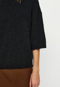 Persona, kas valkā melnu pūkainu īsām piedurknēm svārku un brūnus svārkus, ar redzamu roku un roku uz viendabīga fona.