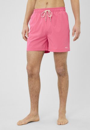 Homme portant un short de bain rose avec un cordon blanc, debout pieds nus devant un fond uni gris clair.