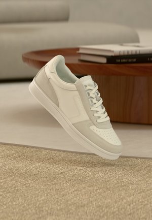 Baskets blanches et gris clair avec lacets, positionnées en l’air au-dessus d’un tapis beige texturé et devant une table en bois avec des livres empilés.