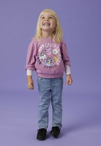 Sudadera lila con caracteres de dibujos animados, texto blanco, mangas enrolladas; combinada con jeans azul claro y zapatos negros. Fondo sencillo.