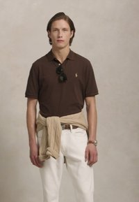 Camisa polo castanha, calças brancas e um pulôver de malha bege claro amarrado à volta da cintura. Óculos de sol pretos e um relógio prateado estão usados.