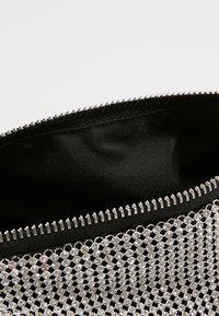 Stříbrná síťovaná kabelka s ozdobnými kamínky a černým textilním interiérem; má zipový uzávěr na vrchu.