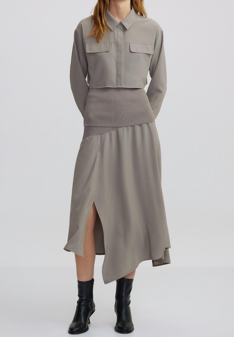 Femme portant un haut cropped à manches longues taupe et une jupe asymétrique assortie avec une fente cuisse, accompagnée de bottines noires à la cheville.