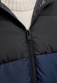 Dunjakke med sort og marineblå blokkfargedesign, med glatt tekstur, full glidelås og ribbet beige genserkrage under.