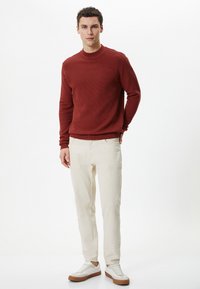 Czerwony sweter o teksturowej powierzchni z okrągłym dekoltem, kremowe spodnie oraz białe sneakersy z beżowymi podeszwami. Prosty design bez wyraźnych wzorów.