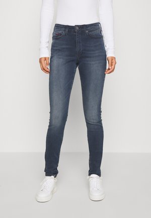Tmavě modré skinny džíny vyrobené z denimu s mírným vyblednutím. Mají pět kapes, zapínání na knoflík a střední pas. Nosí se s bílými teniskami.