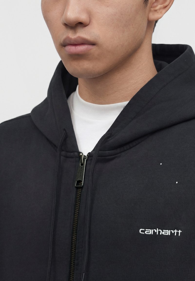 Jeune homme portant un sweat à capuche zippé noir Carhartt par-dessus un t-shirt blanc, montrant la capuche et le logo sur la poitrine.