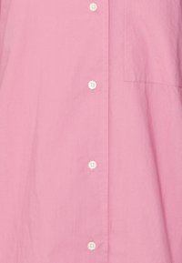 Camicia button-up rosa chiaro con bottoni bianchi, la texture del tessuto appare morbida, include una tasca sul petto e una vestibilità leggermente ampia.