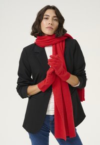 Røde uldhandsker og et matchende ribstrikket tørklæde, parret med en sort blazer og hvid skjorte, vist mod en neutral baggrund.