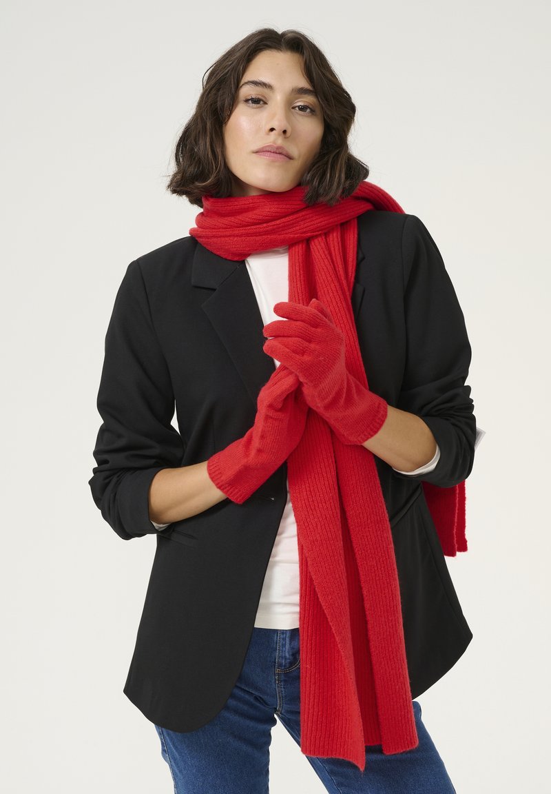 Røde uldhandsker og et matchende ribstrikket tørklæde, parret med en sort blazer og hvid skjorte, vist mod en neutral baggrund.