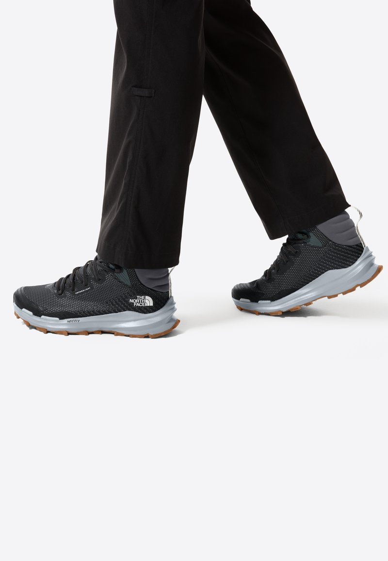 Personne portant des chaussures de trail noires avec des semelles grises et un pantalon noir, marchant sur un fond blanc.