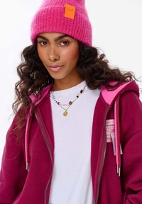 Bordeaux mikina na zip s růžovými akcenty, obsahující přední zip, hluboké kapsy a bílý tričko pod ní. Růžová chlupatá čepice dokončuje vzhled.