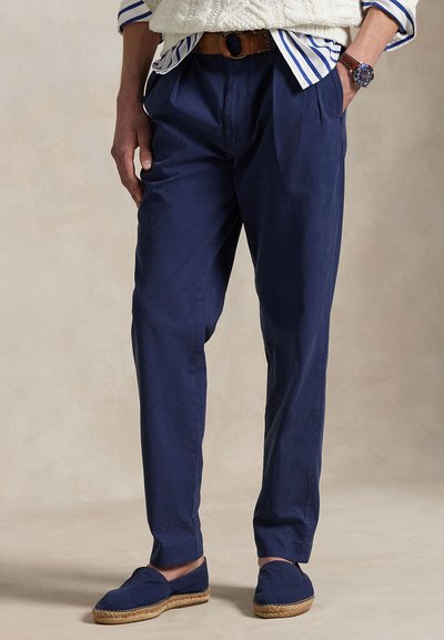 Uomo che indossa pantaloni blu navy, cintura marrone, camicia a righe con maglione bianco, espadrillas blu e orologio da polso, in piedi con le mani in tasca.