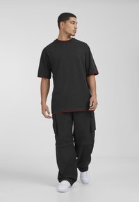 Camiseta negra de gran tamaño con ribete rojo, combinada con pantalones cargo negros que tienen grandes bolsillos y puños elásticos, llevados con zapatillas blancas.