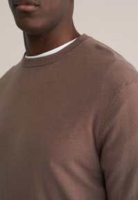 Pull marron tricoté avec un col rond côtelé, une texture lisse et une silhouette ajustée. Superposé sur une chemise blanche.