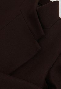 Tela marrón oscuro con una superficie lisa y texturada. Presenta pliegues y costuras, mostrando un diseño estructurado con elementos angulados.