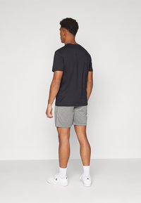 Schwarzes Baumwoll-T-Shirt kombiniert mit grauen gemusterten Shorts. Modell trägt weiße Sneaker und weiße Socken, von hinten gezeigt. Einfaches Design.