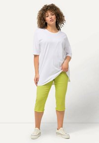 Witte tuniek met korte mouwen, gecombineerd met felgroene cropped pants met rafelige onderranden. Het model draagt witte sneakers, die een casual uitstraling benadrukken.