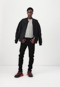 Muž stojaci na bielom pozadí, oblečený v čiernej bomberovej bunde, svetlošedej mikine, čiernych nohaviciach a čiernych a červených teniskách.