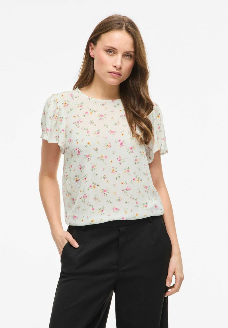 Femme aux longs cheveux bruns portant un chemisier blanc à manches courtes à motifs fleuris et un pantalon noir, debout avec une main dans la poche.