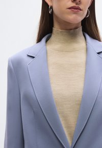 Σιέλ σακάκι με κομμένες γιακάδες φορεμένο πάνω από μπεζ ζιβάγκο. Ορατά ασημένια κρίκοι σκουλαρίκια. Υφασμάτινη υφή, εφαρμοστή σχεδία�ση.