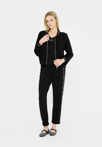 Zwart sportkledingset met een jas met ritssluiting en logodetails, en bijpassende broek met logostrepen aan de zijkant, gemaakt van een zachte, rekbare stof.