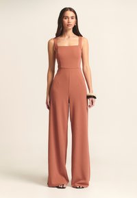Roodbruine jumpsuit met een vierkante halslijn, brede straps en wijde broekspijpen. Gemaakt van een gladde stof en voorzien van zijzakken.