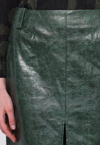 Zelená sukně z umělé kůže s vrásčitým texturou, vybavená předním rozparkem a zajištěným pasem. Detailní pohled s rukou pro měřítko.