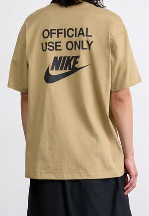 Osoba nosząca beżową koszulkę Nike z napisem "TYLKO DO UŻYTKU OFICJALNEGO" oraz czarne logo Nike na plecach, w połączeniu z czarnymi spodniami.