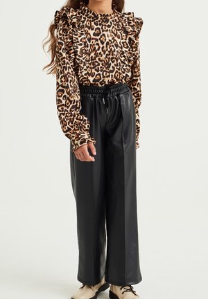 Junge Person trägt eine Bluse mit Leopardenmuster und Rüschen, Knöpfe, schwarze, weit geschnittene Hosen aus Kunstleder und beige Schnürschuhe.