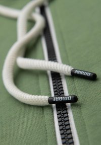 Grønt stof med en tekstureret overflade, prydet med en sort lynlås og hvide snore med sorte plastikspidser, mærket "PUMA".