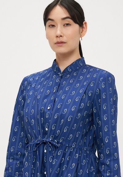 Μπλε φόρεμα με σχέδια, που διαθέτει ψηλό γιακά, κούμπωμα με κουμπιά και ζώνη με κορδόνι στη μέση. Το ύφασμα έχει λεία υφή με αντίθετες λευκές λεπτομέρειες.