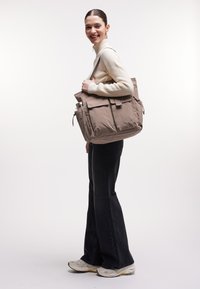 Sac fourre-tout en nylon beige avec deux poches avant, des bretelles ajustables et une poche latérale pour une bouteille d'eau. Texture lisse et conception robuste.