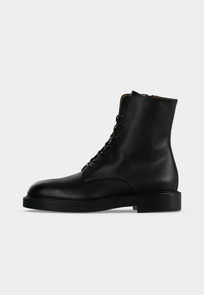 Μαύρο δερμάτινο μποτάκι αστραγάλου με κορδόνια, επίπεδη σόλα και στρογγυλεμένη μύτη, εμφανιζόμενο από το πλάι σε απλό φωτεινό φόντο.