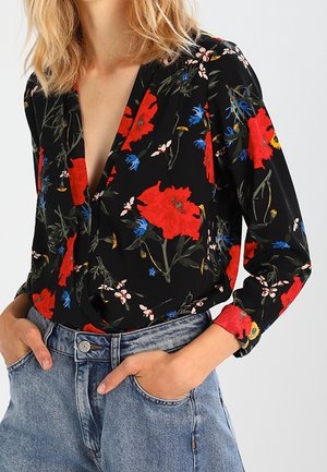 Woman wearing black blouse with red and blue floral print, tucked into light blue high-waisted jeans, hands in pockets.