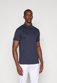 Polo azul-escuro em tecido respirável com colarinho, abertura de três botões e detalhe de logótipo na gola, combinado com calças brancas.