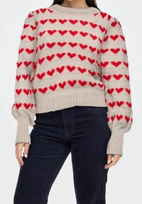 Beżowy sweter z czerwonymi wzorami serc, ściągaczami na rękawach i dole, luźny fason, dzianinowa faktura, miękki materiał. Noszony z ciemnymi spodniami.