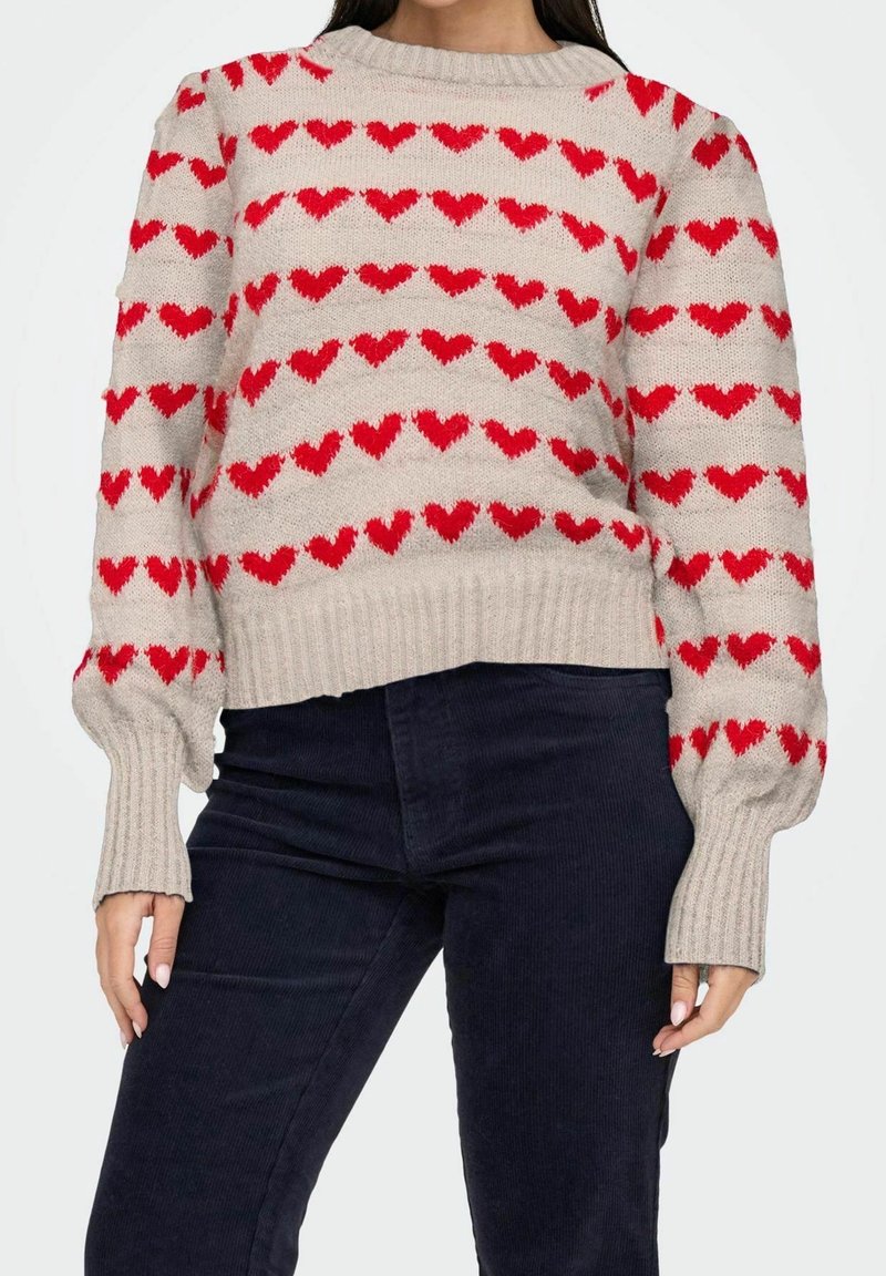 Beżowy sweter z czerwonymi wzorami serc, ściągaczami na rękawach i dole, luźny fason, dzianinowa faktura, miękki materiał. Noszony z ciemnymi spodniami.