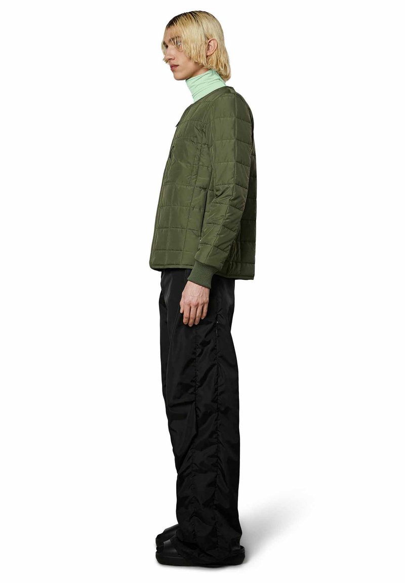 Giacca trapuntata verde oliva con scollatura a girocollo e polsini a costine, abbinata a pantaloni neri con vestibilità rilassata e una leggera texture.