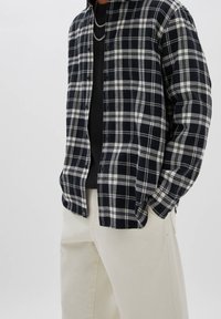 Uomo con camicia a quadri bianca e nera sopra una maglietta nera con catene d'argento, abbinata a pantaloni color crema, con le mani nelle tasche.