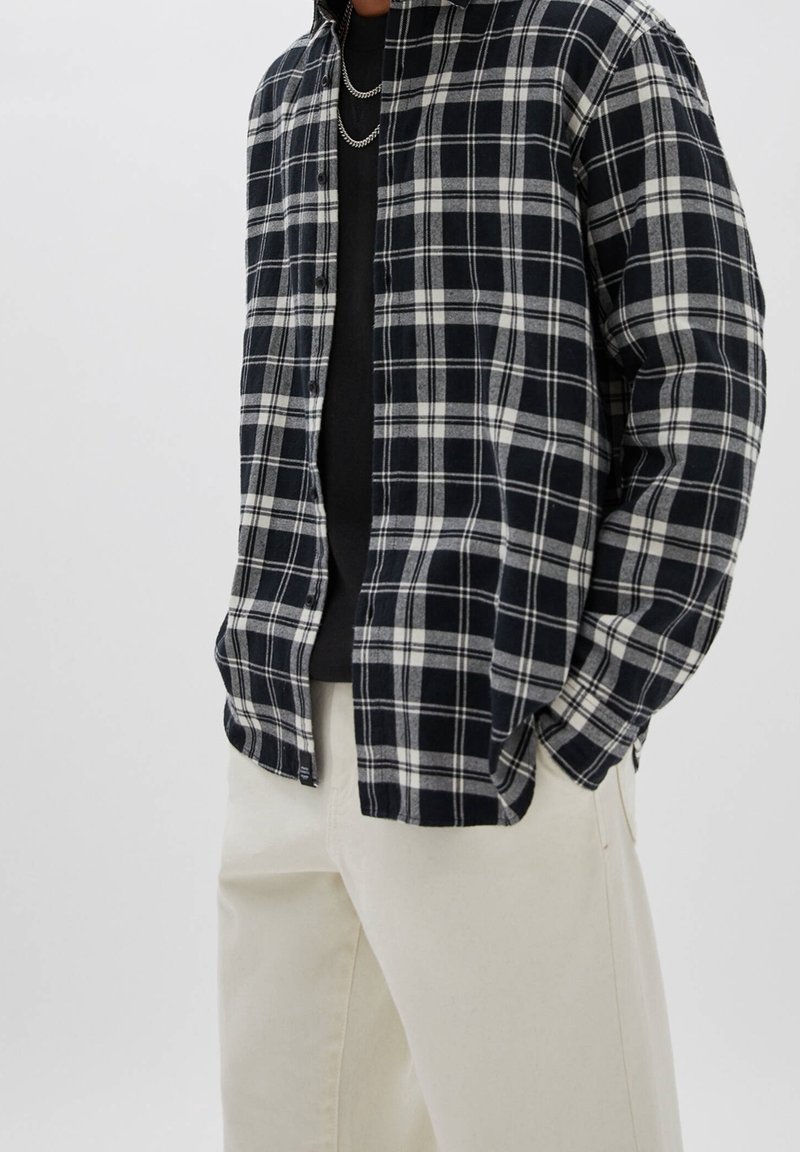 Uomo con camicia a quadri bianca e nera sopra una maglietta nera con catene d'argento, abbinata a pantaloni color crema, con le mani nelle tasche.